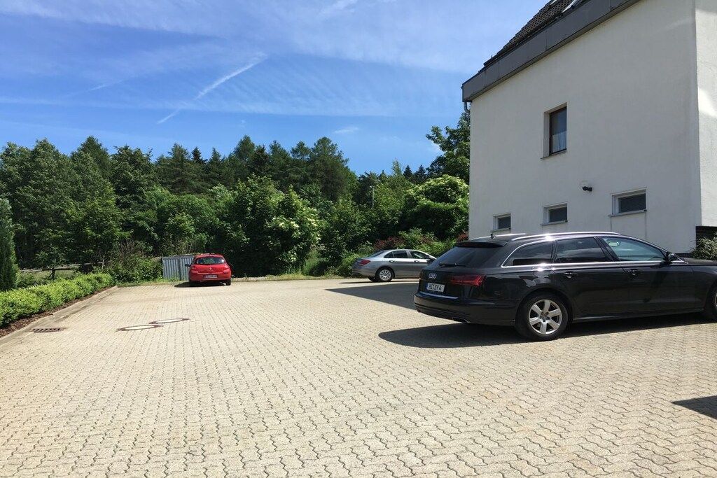 Un camino de entrada pavimentado que conduce a un edificio de fachada blanca, con coches aparcados y árboles al fondo.