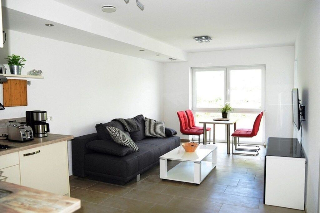 Ein moderner Wohnbereich mit Sofa, Couchtisch und roten Sesseln. Es gibt eine Küchenzeile mit Toaster und einer Pflanze.
