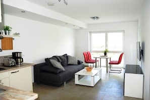 Ein moderner Wohnbereich mit Sofa, Couchtisch und roten Sesseln. Es gibt eine Küchenzeile mit Toaster und einer Pflanze.