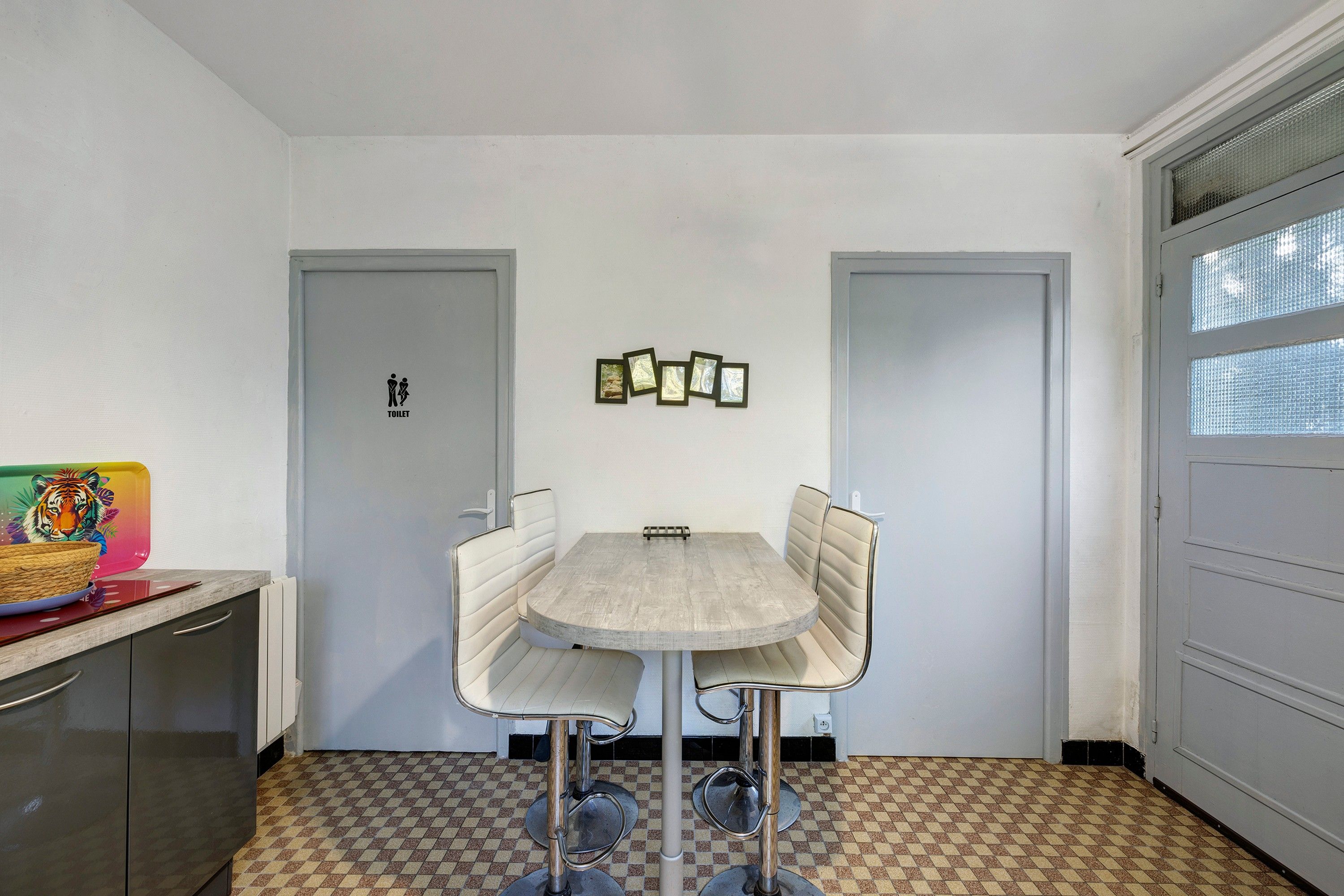 Une petite pièce fermée, avec une table ronde et quatre chaises, un radiateur, une porte portant un panneau indiquant les toilettes et une fenêtre avec un motif en croisillons.