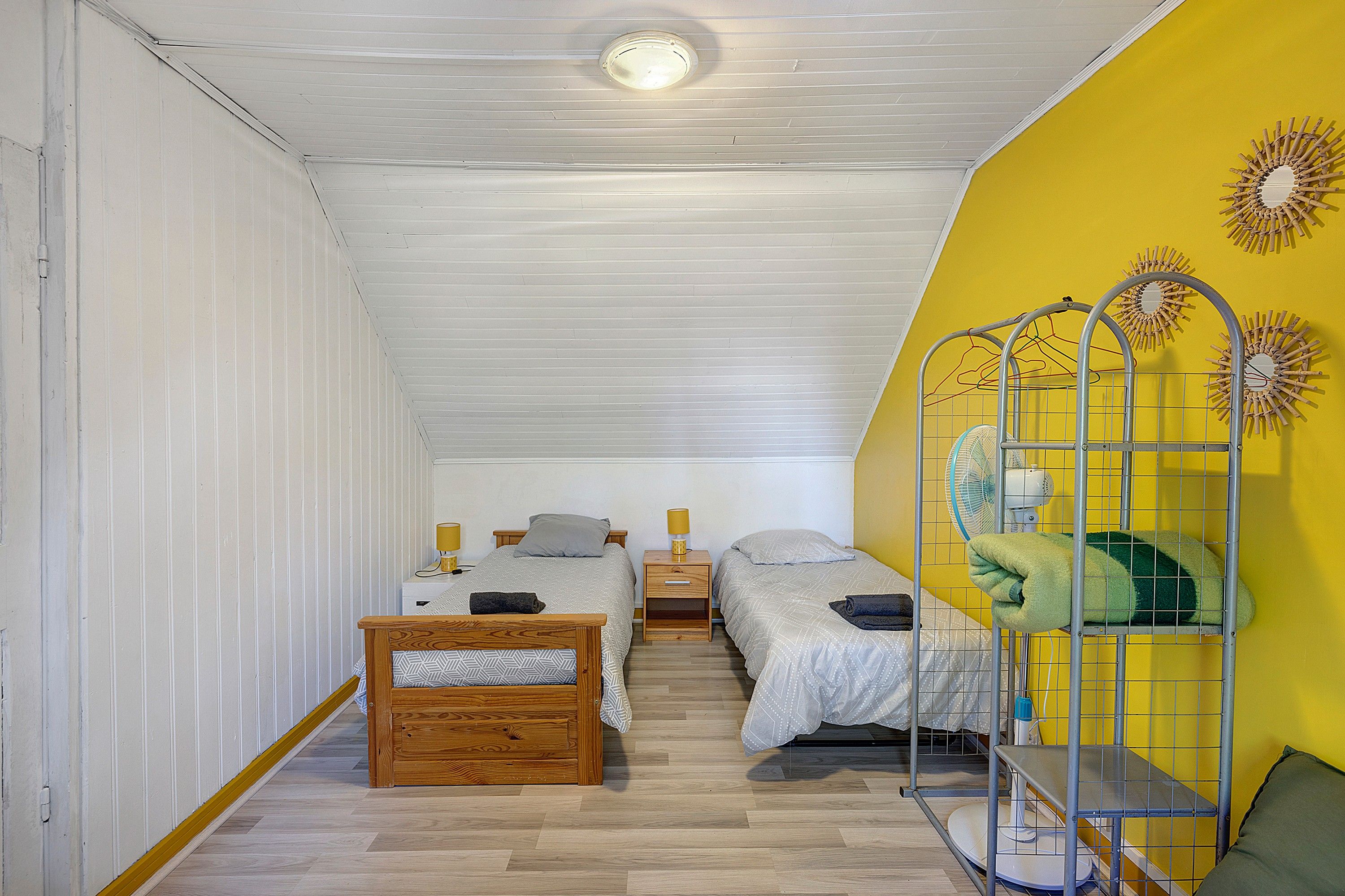 Una habitación con dos camas, piso de madera, pared amarilla y una lámpara en el techo.