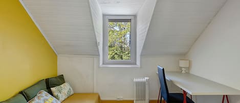 Une chambre avec un lit, un bureau, une chaise, un radiateur et une fenêtre donnant sur des arbres.