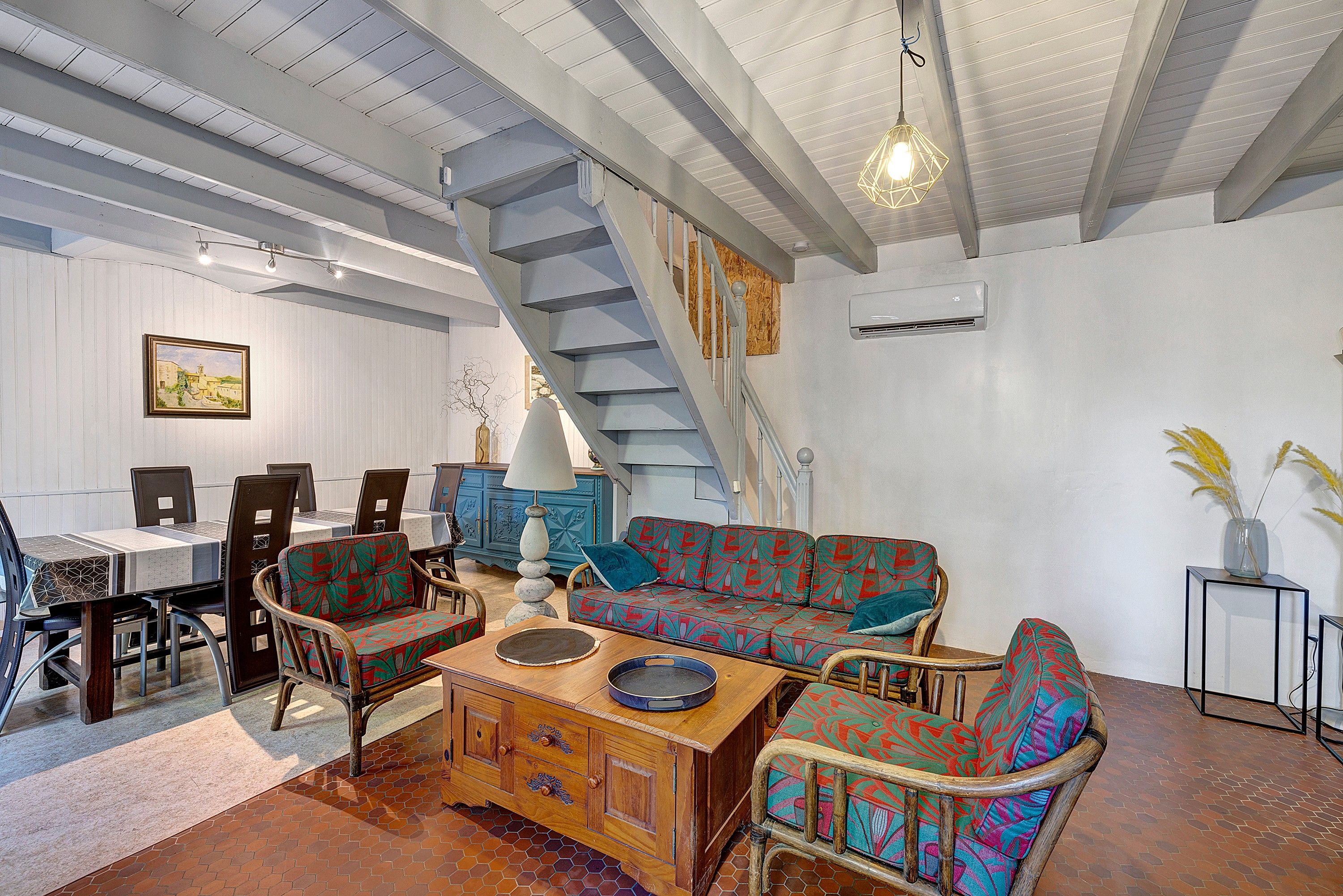 A spacious living area with a wooden coffee table, patterned sofa, and a dining table with chairs. A staircase with a wooden railing leads to an upper level.