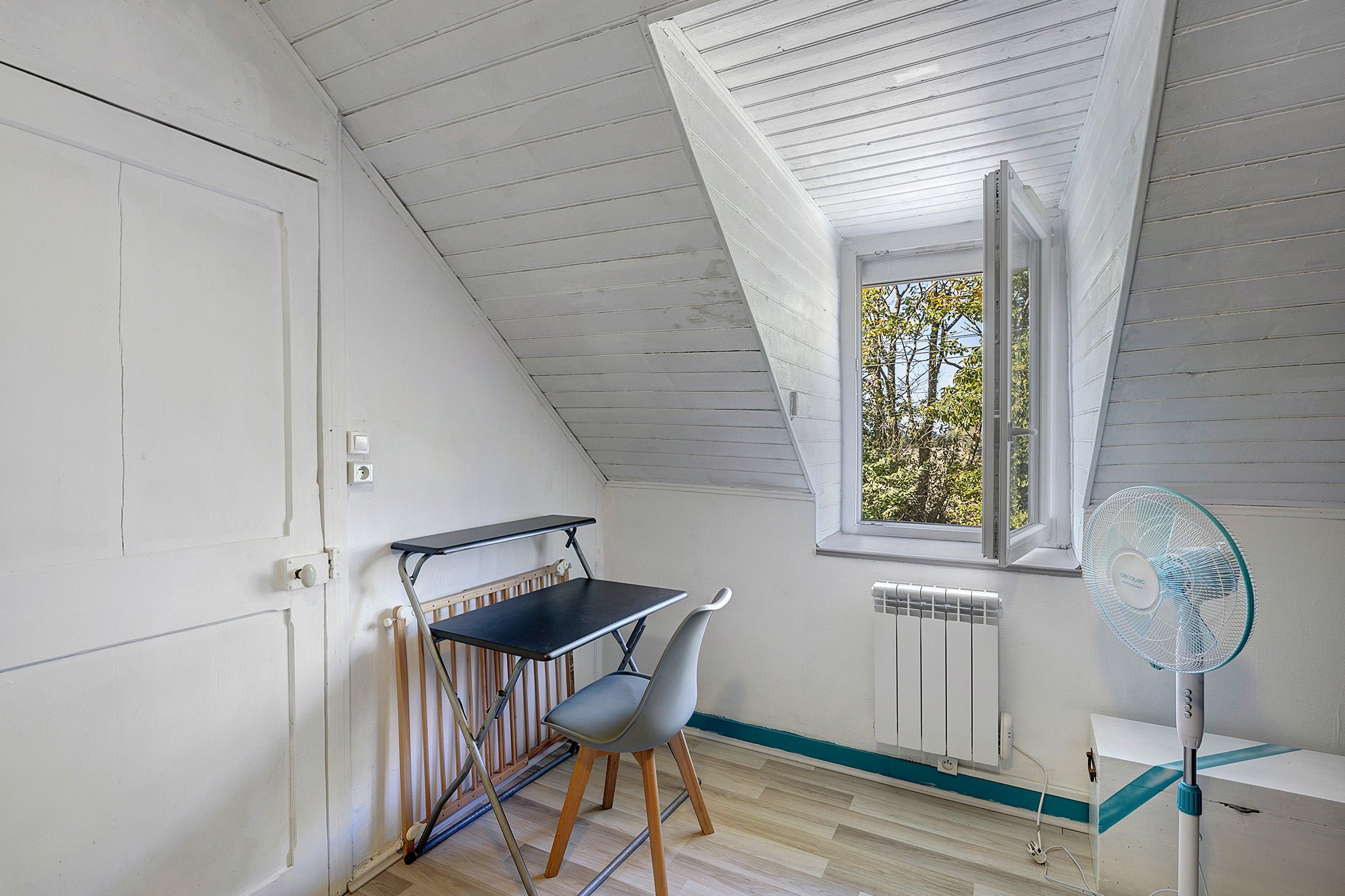 A small attic room with a slanted ceiling, a window with a view of trees, a desk with a chair, and a standing fan.