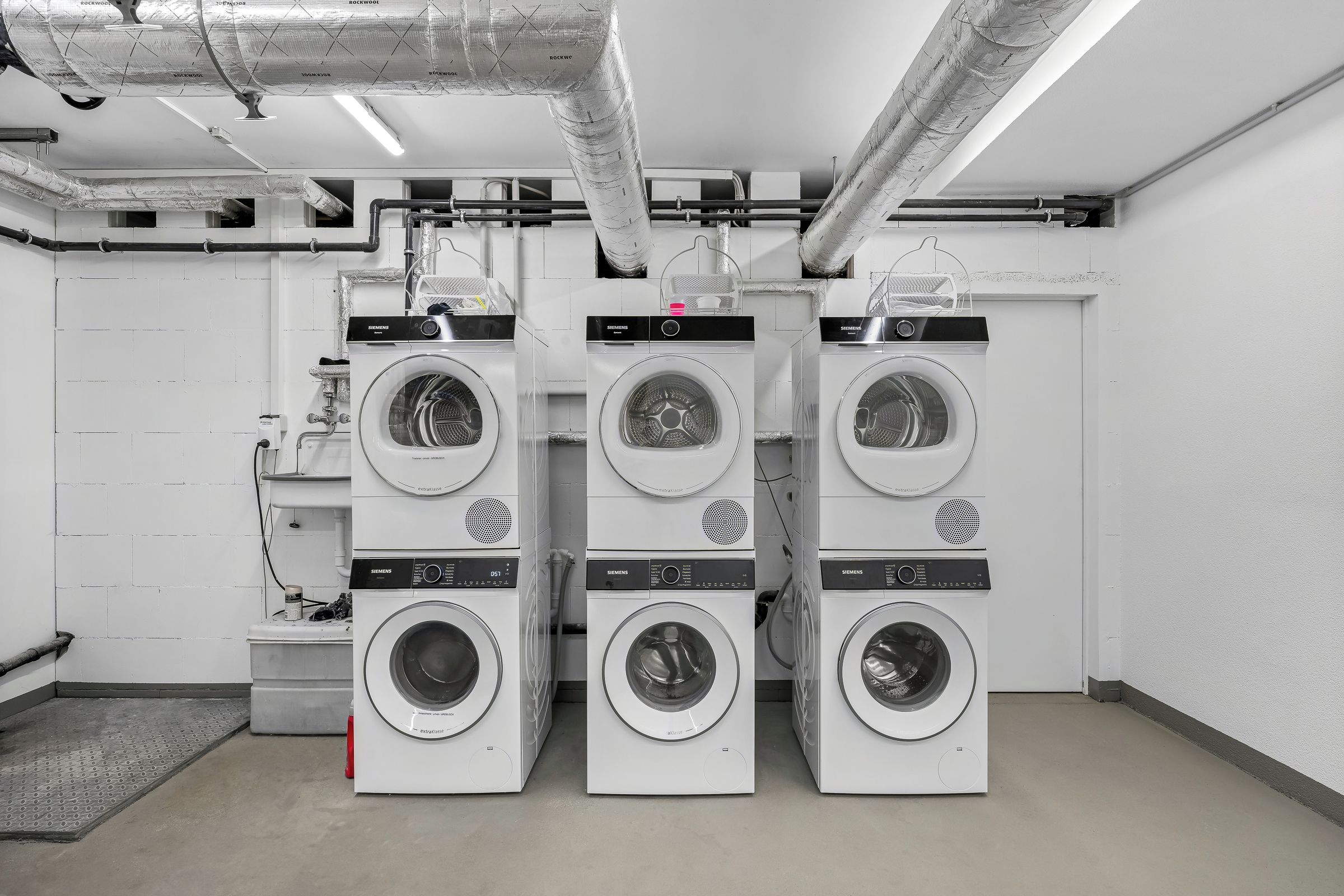 Une laverie équipée de plusieurs machines à laver de la marque Siemens, de conduits de ventilation et d’un sol carrelé.