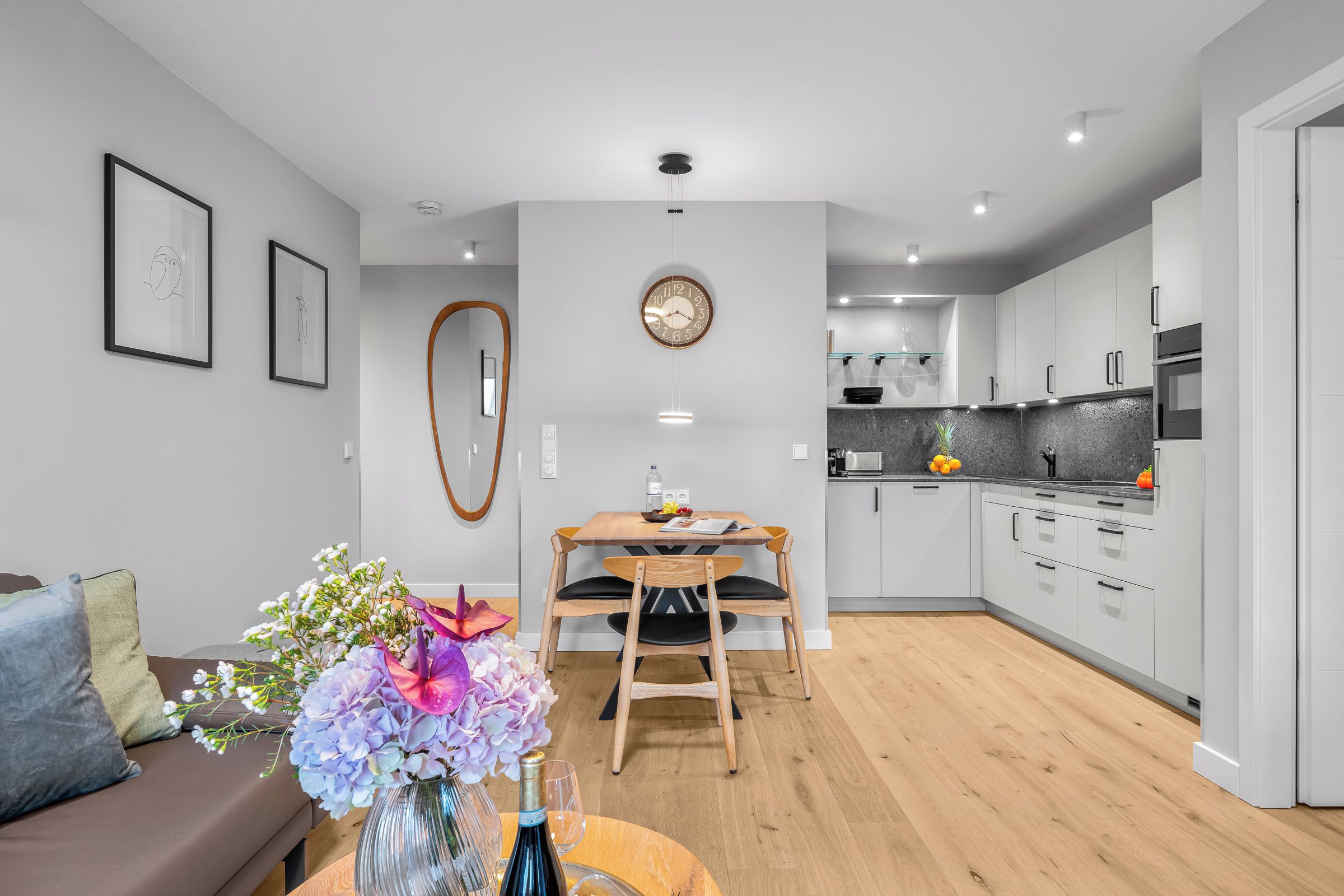 A modern kitchen with white cabinets, a dining area with a wooden table and chairs, and a living room with a brown sofa and a vase of flowers.