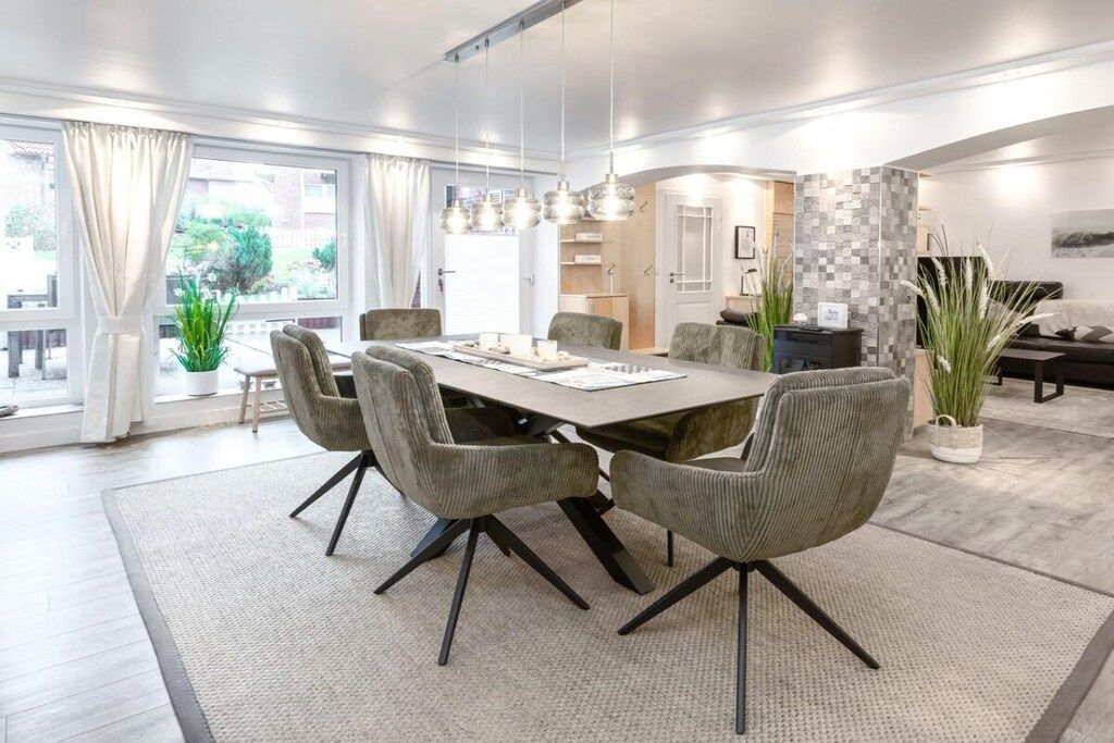 A modern dining area with a rectangular table, six upholstered chairs, pendant lighting, and a view of the outdoors through large windows.