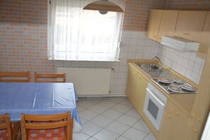 Pieni keittiö, jossa on laatoitettu lattia, ikkunassa verhot, pöytä tuoleineen ja puiset kaapit.