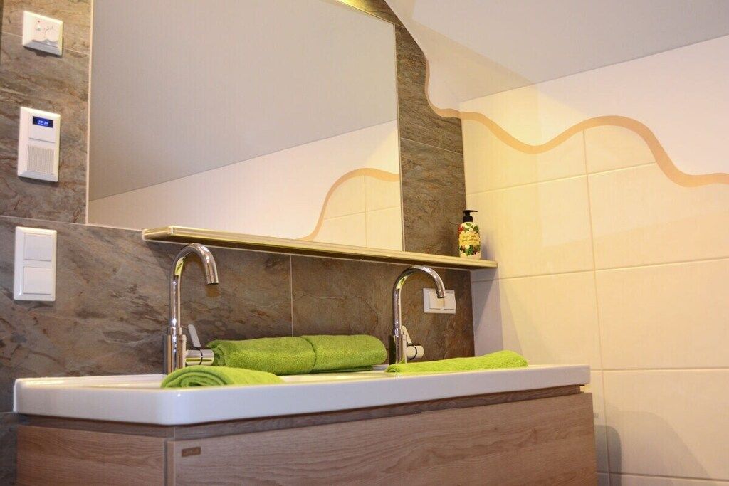A bathroom with a white sink, two faucets, a green towel, and a soap dispenser on a shelf.