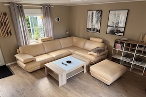 A modern living room with a beige sectional sofa, a white coffee table, and a matching ottoman. There is a bookshelf with books and decorative items. - Holiday home with 2 bedrooms (Joachimsthal)
