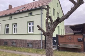 En tvåvåningsbyggnad med en fasad av tegelsten i botten och en grön överdel, ett rött tegeltak och ett träd i förgrunden.