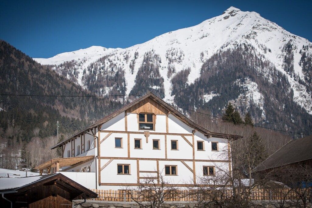A traditional chalet with a symmetrical facade, multiple windows, and a wooden roof, set against a backdrop of snow-covered mountains.
