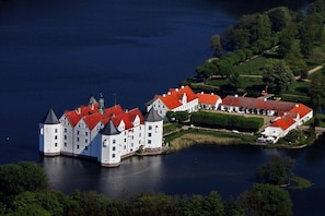 Eine große, weiße Burg mit roten Dächern, gelegen an einem See, umgeben von viel Grün.
