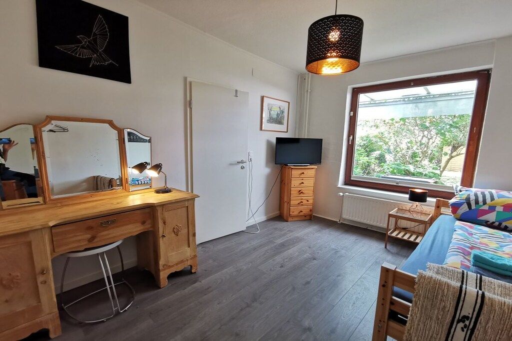 Un dormitorio con un escritorio de madera, un televisor, una cama con una colcha colorida, una ventana con vistas a la vegetación y una lámpara colgante.