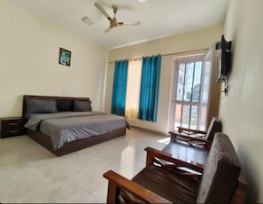 A bedroom with a bed, a nightstand, a desk, a chair, a ceiling fan, and a window with curtains. - Hotel Gliders Inn (Baijnath)
