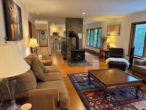 A cozy living room with a fireplace, a sofa, a coffee table, and a view of the kitchen in the background. - Luxury Chalet on 1.5 Acres: Mountain Views, Hot Tub, Gym, fire pit & Ping-Pong (Windham)