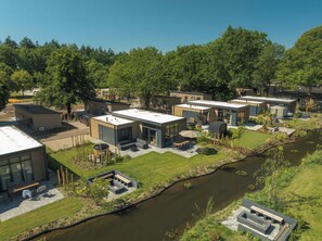 A modern residential complex with multiple houses, a central courtyard, and a small canal. - Stylish 6-person home with three bedrooms, a modern kitchen, air conditioning, a cozy outdoor space with a luxury pergola, private hot tub, sauna and a lounge area with an outdoor fireplace. Enjoy comfort and nature in the Veluwe. (Lieren)