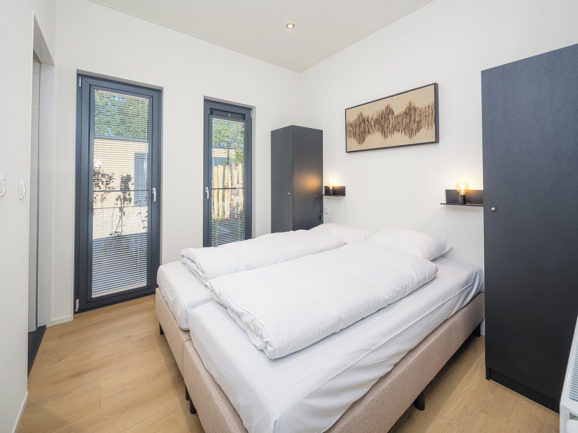 Ein modernes Schlafzimmer mit einem großen Bett, zwei Türen, einem Gemälde und einem kleinen Tisch mit einer Lampe.