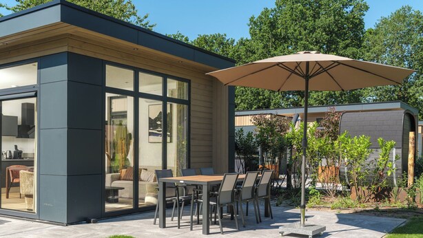 A modern outdoor patio with a dining table and chairs, a large umbrella, and a glass-enclosed kitchen area. - Stylish 6-person home with three bedrooms, a modern kitchen, air conditioning, a cozy outdoor space with a luxury pergola, private hot tub, sauna and a lounge area with an outdoor fireplace. Enjoy comfort and nature in the Veluwe. (Lieren)