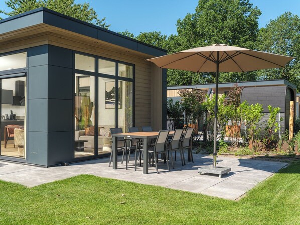 Um pátio moderno ao ar livre com mesa e cadeiras, um guarda-sol grande e uma área de cozinha envidraçada.