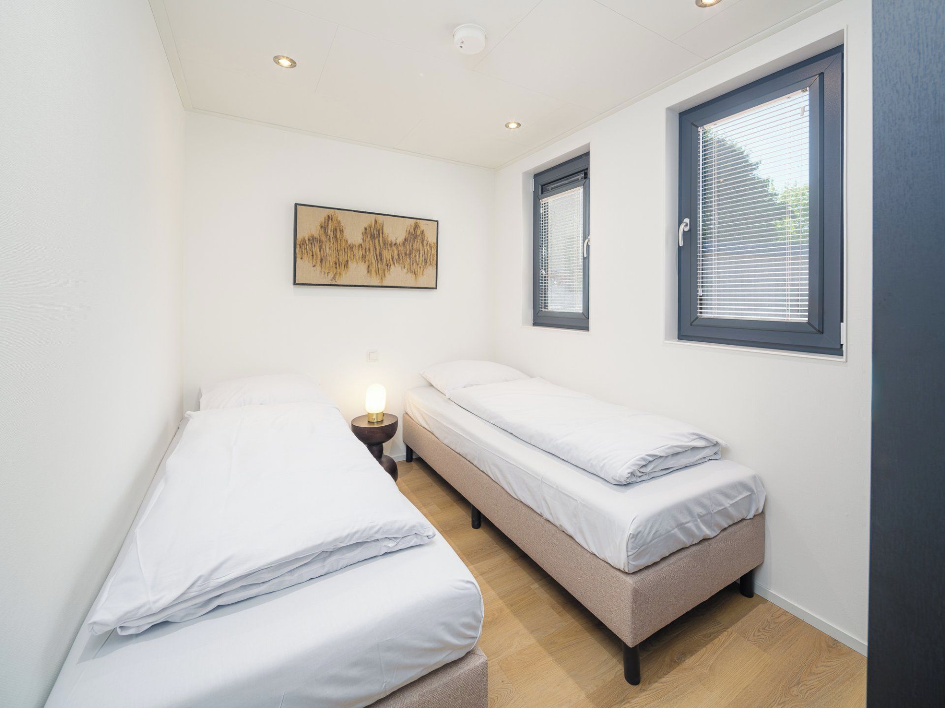 Un dormitorio pequeño y luminoso con dos camas individuales, una mesita de noche con lámpara, una obra de arte colgada en la pared y una ventana con persianas.