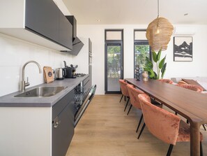 A modern kitchen with a built-in dishwasher, a sink, and a stove. There is a dining area with a wooden table and chairs. - Stylish 6-person home with three bedrooms, a modern kitchen, air conditioning, a cozy outdoor space with a luxury pergola, private hot tub, sauna and a lounge area with an outdoor fireplace. Enjoy comfort and nature in the Veluwe. (Lieren)