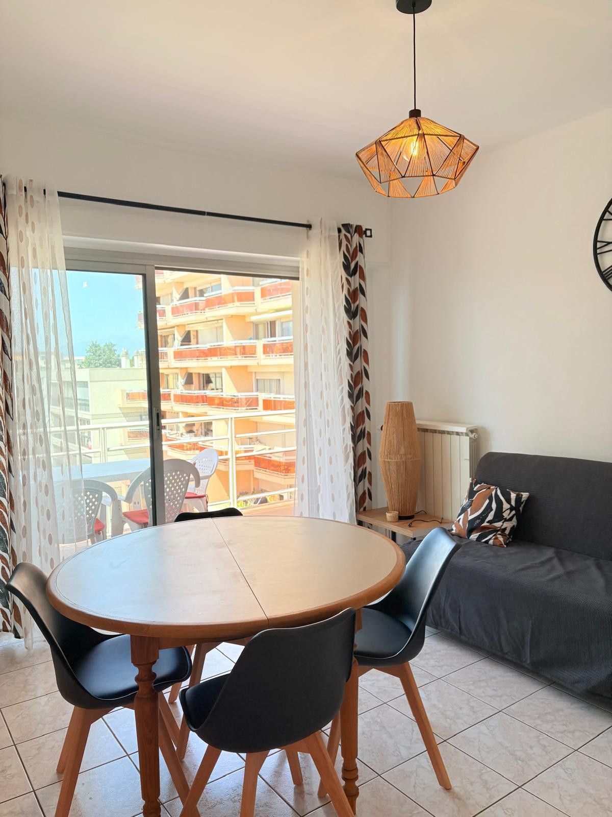 A modern living room with a round wooden table, two black chairs, a grey sofa, and a patterned throw pillow. A unique geometric pendant light hangs from the ceiling.