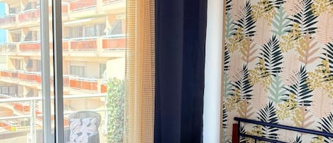 A hotel room with a bed, a view of a building through a window, and a balcony with chairs.