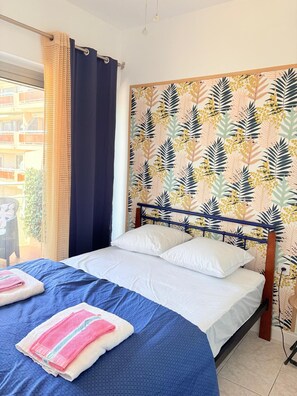 A hotel room with a bed, two pillows, a blue bedspread, a pair of towels, a window with a curtain, and a wall with a leaf pattern. - Superb apartment on the port of St Cyprien (Saint-Cyprien)