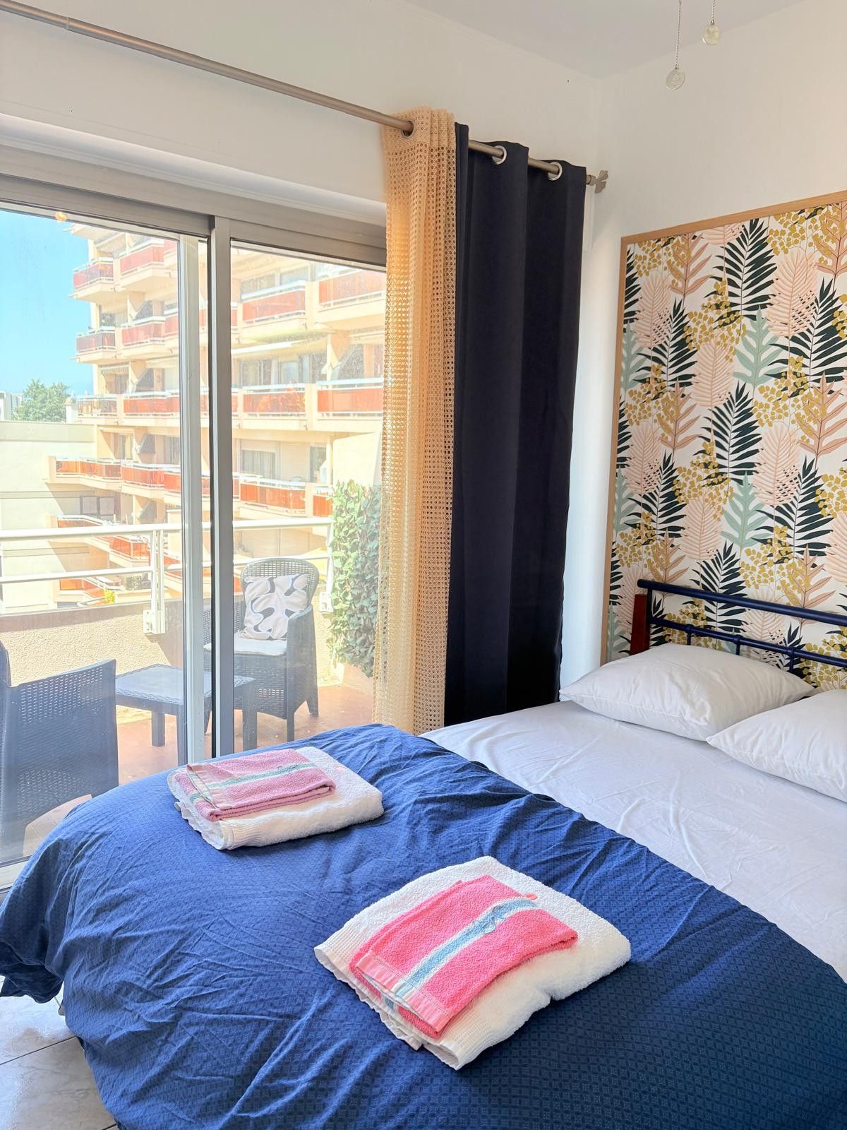 A hotel room with a bed, a view of a building with balconies, and a chair outside.