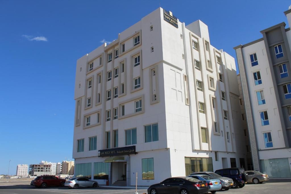 Um edifício de hotel com vários andares e fachada moderna, com vários espaços de estacionamento e um céu azul claro ao fundo.