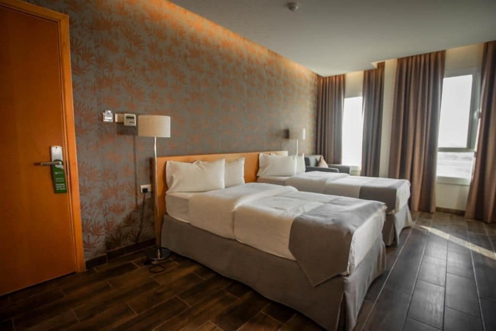 Quarto de hotel com duas camas, piso de madeira, parede estampada e janela com cortinas.