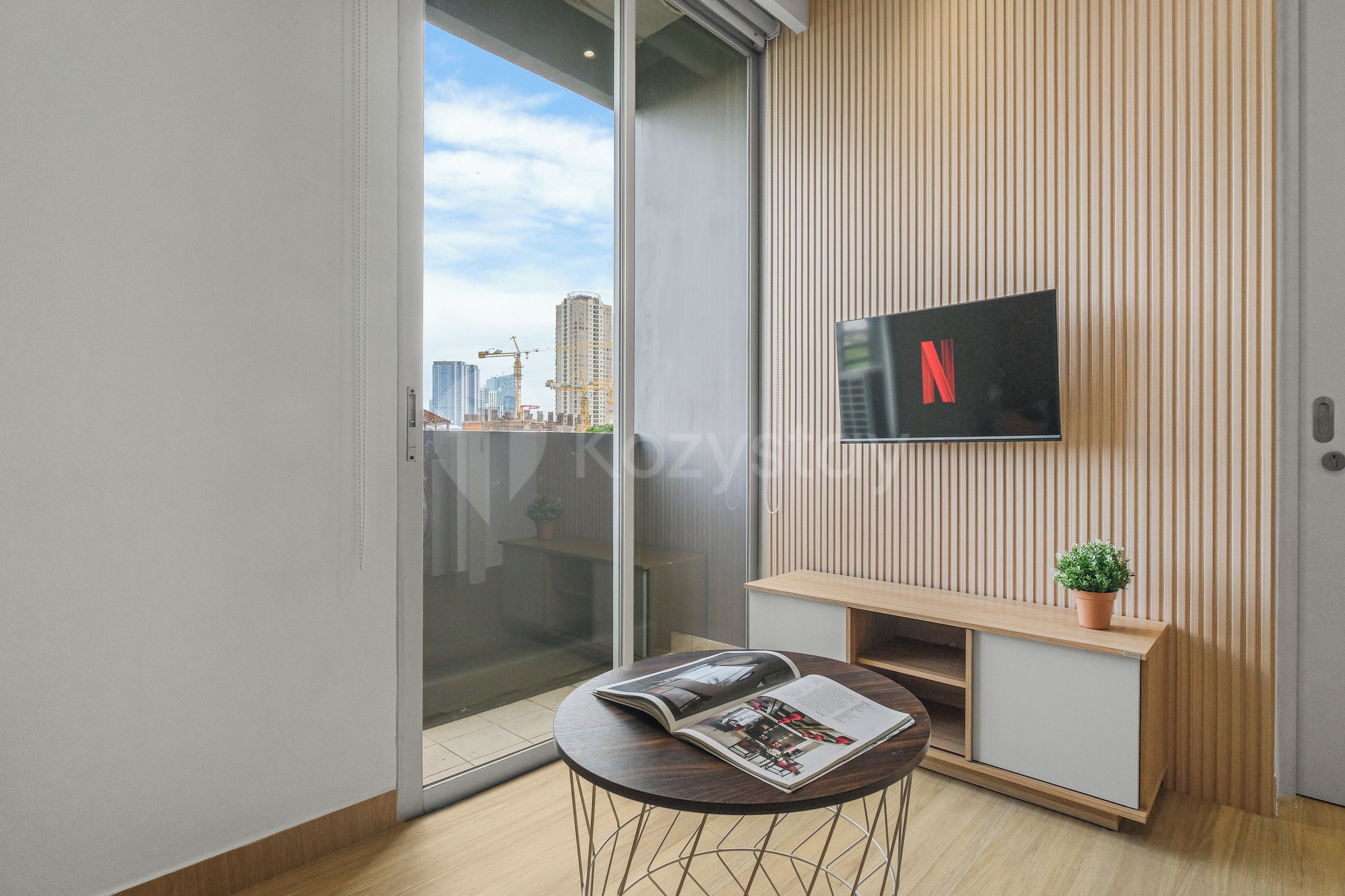 Een moderne woonkamer met een glazen deur naar een balkon, een flatscreen televisie bevestigd aan een houten wandpaneel, een klein rond tafeltje met een tijdschrift en een potplant.