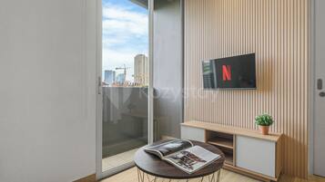 Een moderne woonkamer met een glazen deur naar een balkon, een flatscreen televisie bevestigd aan een houten wandpaneel, een klein rond tafeltje met een tijdschrift en een potplant.