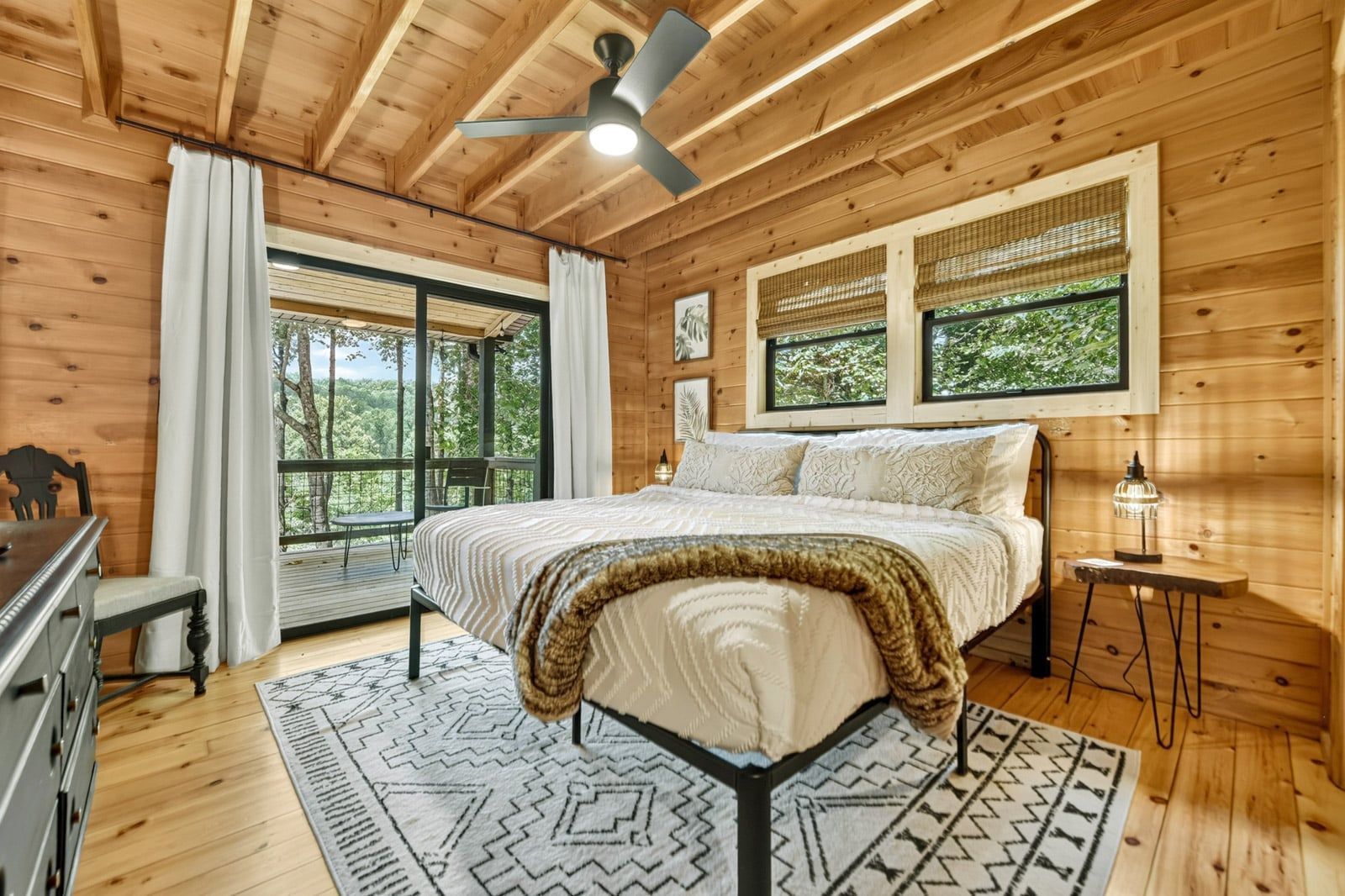 A wooden cabin bedroom with a bed, a ceiling fan, a window with a view, and a small table with a lamp.