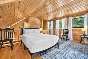 Ein Schlafzimmer einer Holzhütte mit einem Bett, zwei Stühlen, einem Nachttisch und einem Fenster mit Vorhängen.