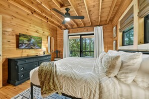 Een slaapkamer in een houten blokhut met een bed, een dressoir, een televisie en een raam met gordijnen.