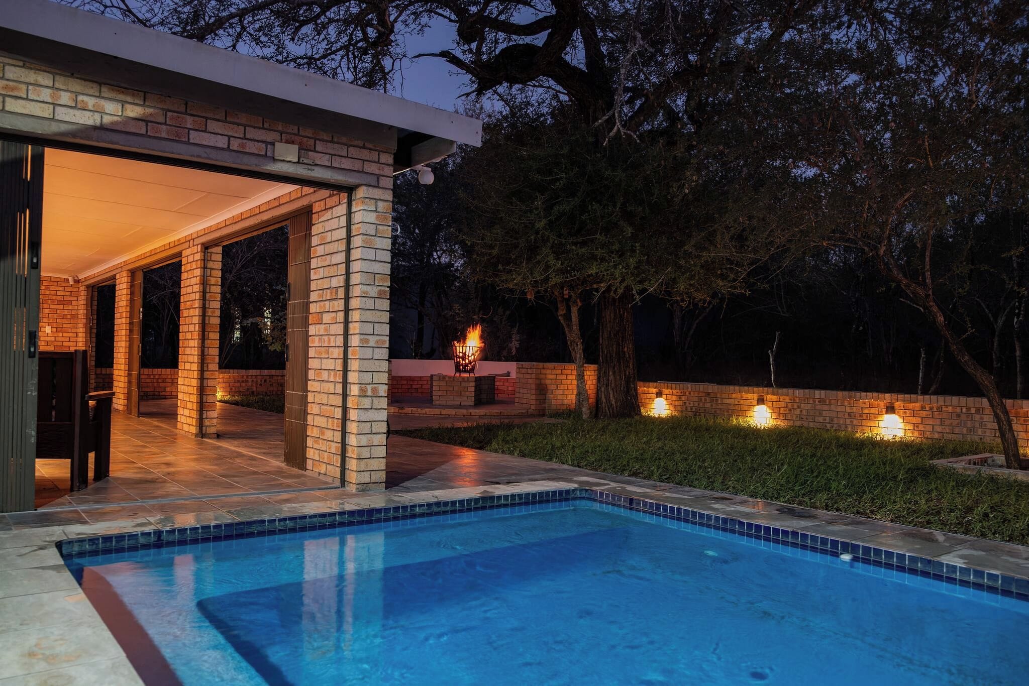 A modern outdoor pool area with a brick patio, a fire feature, and a well-lit entrance.