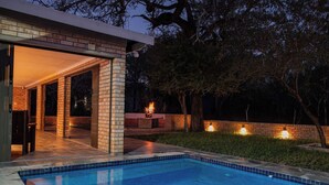 A modern outdoor pool area with a brick patio, a fire feature, and a well-lit entrance. - Kruger Villa - Where Luxury Meets Nature (Nkomazi)