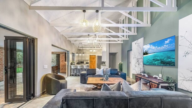 A modern living room with a large sectional sofa, a dining area, and a kitchen in the background. - Kruger Villa - Where Luxury Meets Nature (Nkomazi)