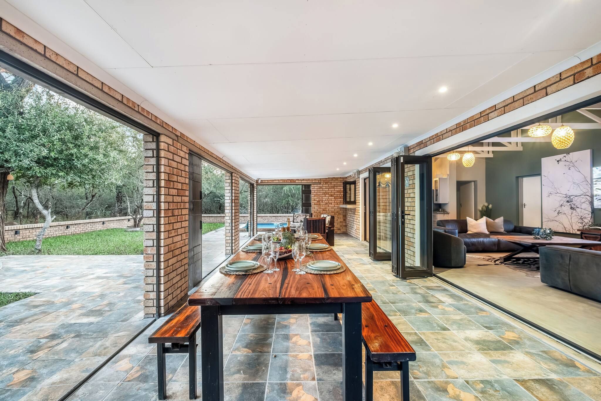 Une vaste salle à manger avec une longue table en bois dressée pour le repas, entourée de portes vitrées donnant sur une piscine et un salon.