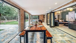 A spacious dining area with a long wooden table set for a meal, surrounded by glass doors leading to a pool and a living area. - Kruger Villa - Where Luxury Meets Nature (Nkomazi)
