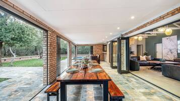 Une vaste salle à manger avec une longue table en bois dressée pour le repas, entourée de portes vitrées donnant sur une piscine et un salon.