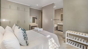 A bedroom with a bed, bedside tables, a wardrobe, a bathroom with a sink and mirror, and a crib. - Kruger Villa - Where Luxury Meets Nature (Nkomazi)