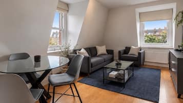 A modern living room with a glass dining table, two sofas, a coffee table, and a view of buildings through large windows.