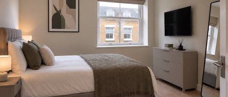 A bedroom with a bed, bedside table, lamp, dresser, TV, and a window with a view of buildings.