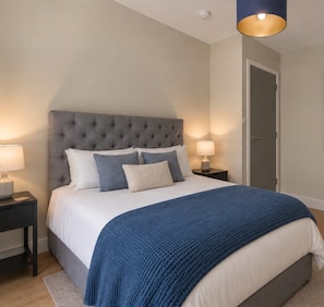 A bedroom with a tufted headboard, a bed with white and blue bedding, two bedside lamps, and a door. - Soho City Canvas • Stylish 2BR • Sleeps 4 (London)