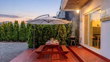 An outdoor patio with a wooden table and benches, a barbecue grill, and a large umbrella.