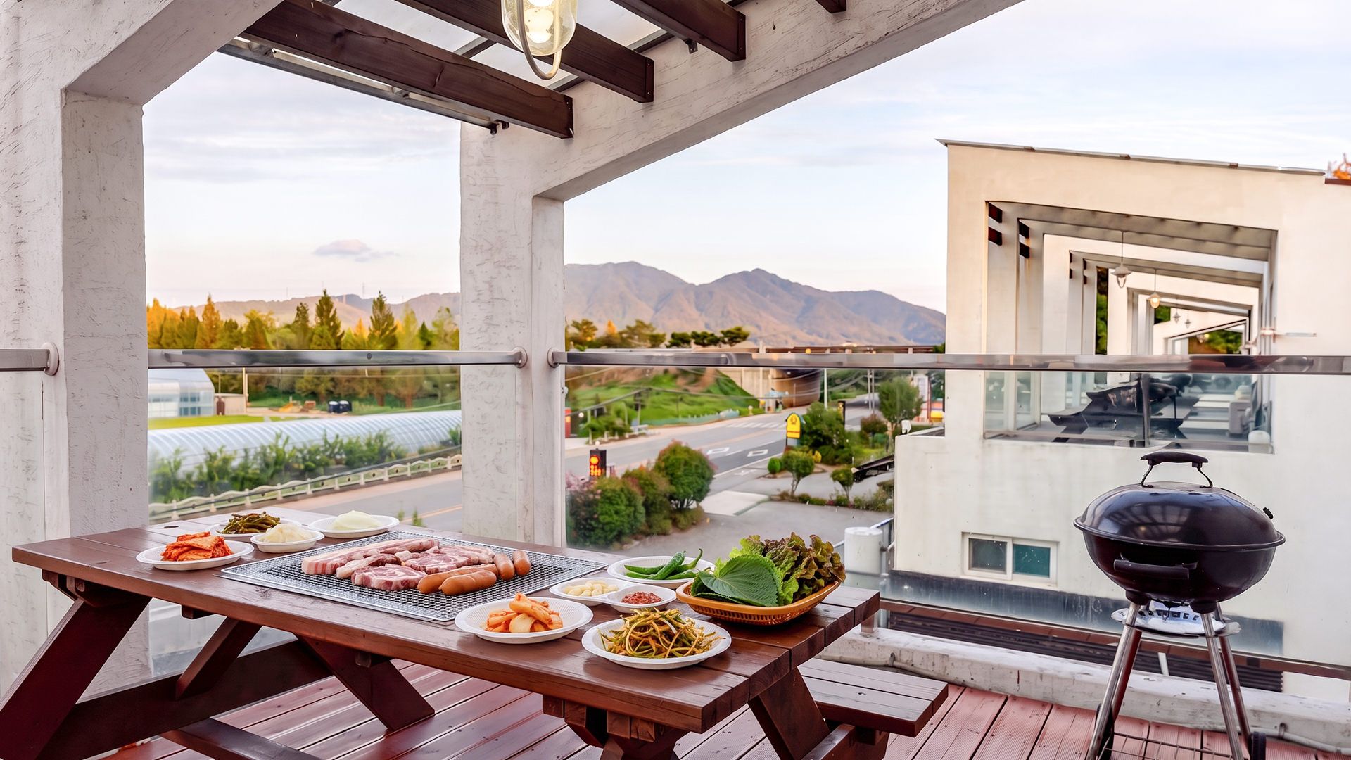 A balcony with a wooden table set for a meal, a barbecue grill, and a view of a cityscape with mountains in the distance.