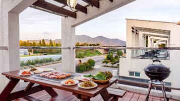 A balcony with a wooden table set for a meal, a barbecue grill, and a view of a cityscape with mountains in the distance.